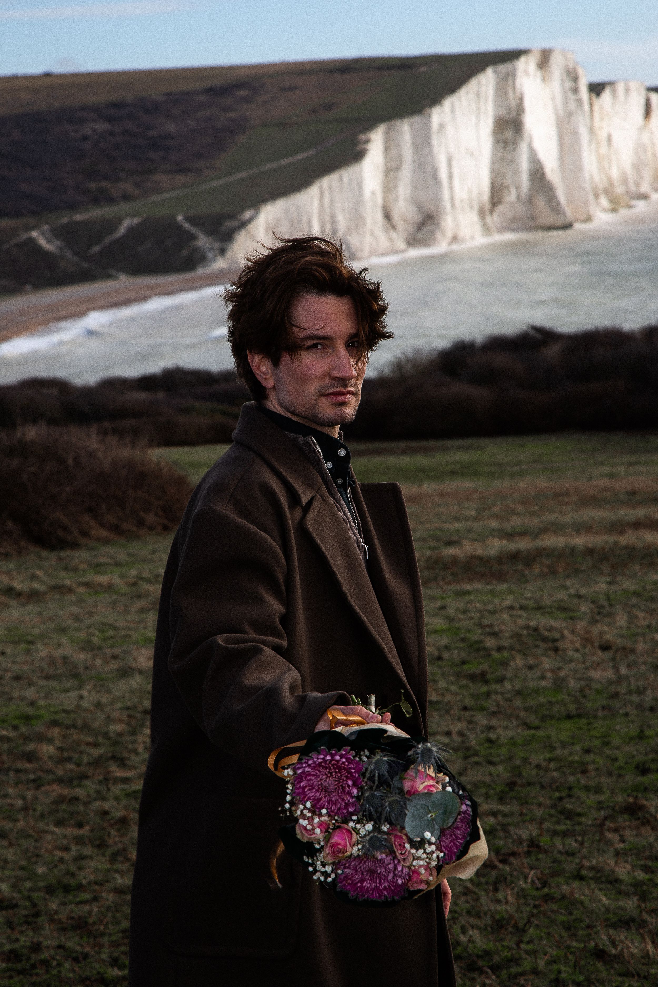 Person in brown coat stands against dramatic white cliffs holding vibrant purple and pink wildflowers.