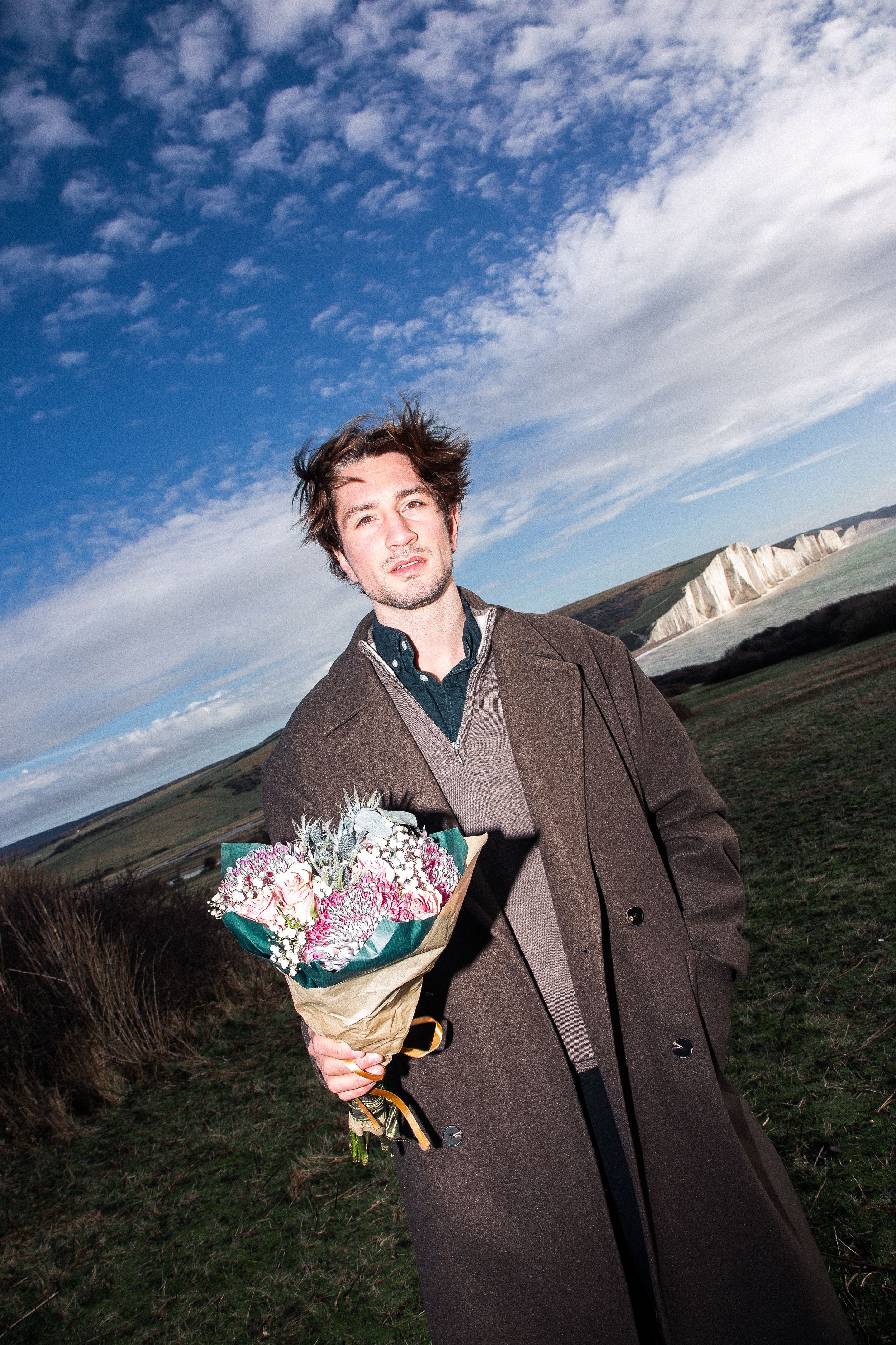 A figure in a brown coat holds a colorful bouquet against a cloudy sky and grassy coastal landscape.