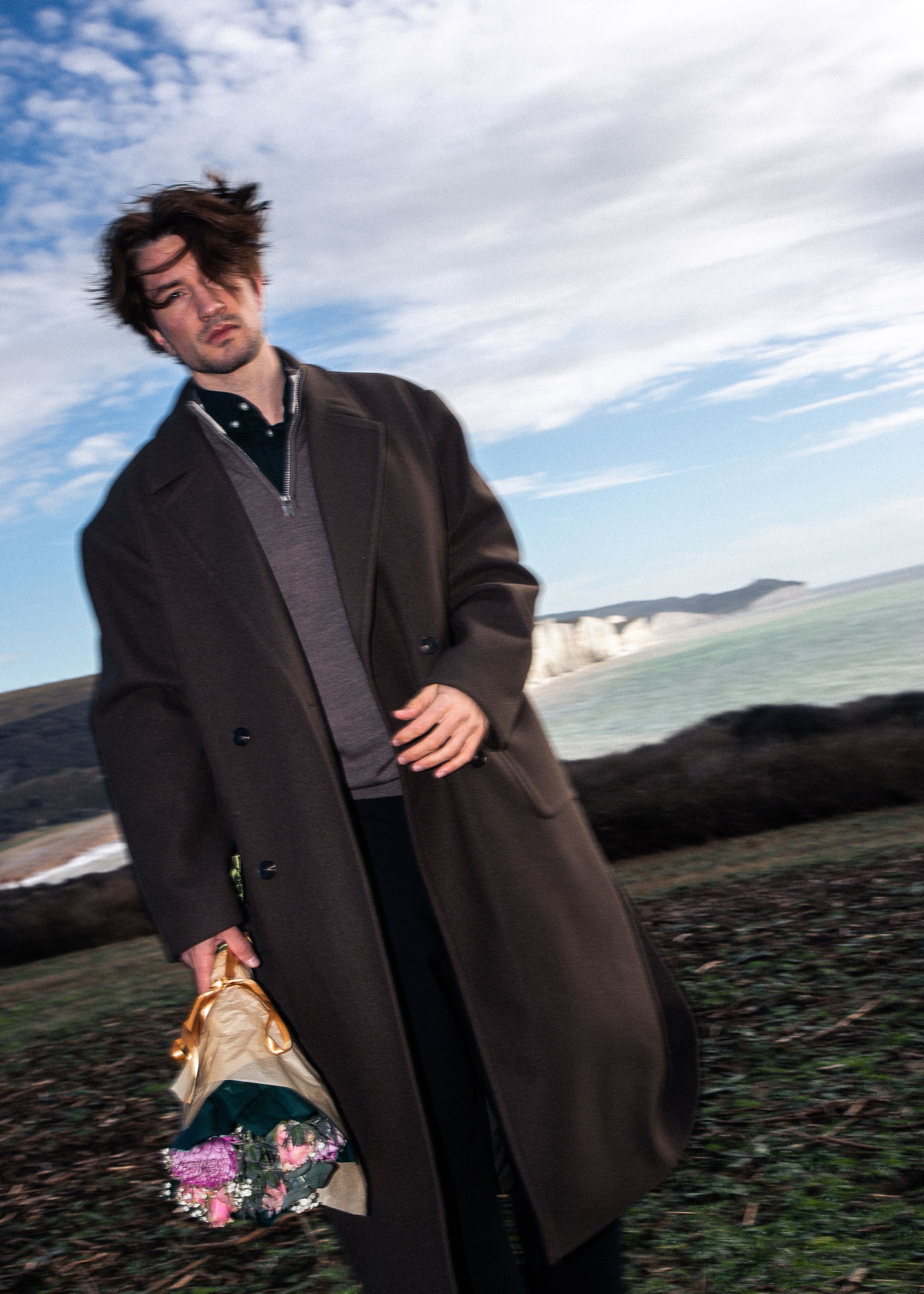 A person in a dark coat walks outdoors on a cloudy day carrying colorful shopping bags against a scenic landscape.
