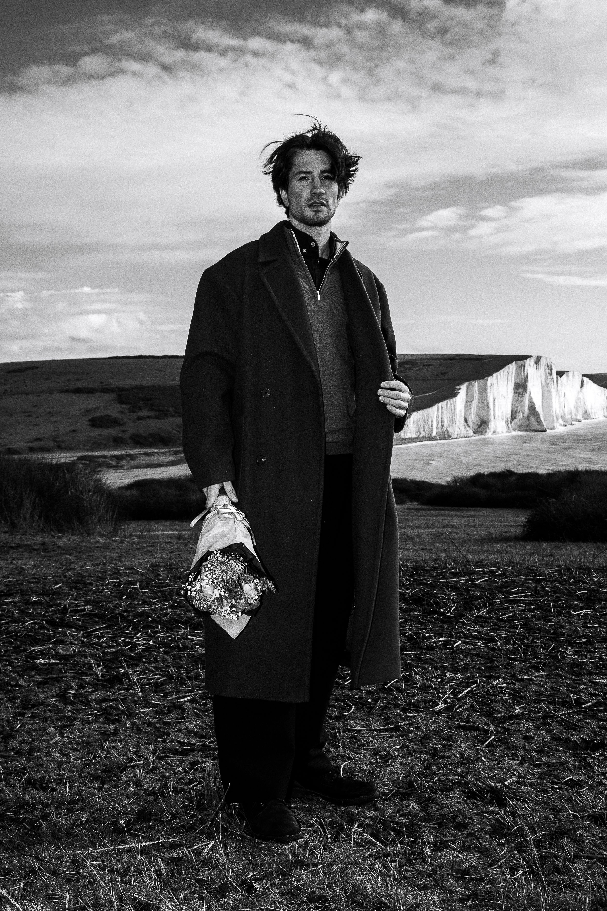 A figure in a dark coat stands on a dramatic coastal landscape with white cliffs in black and white photography.
