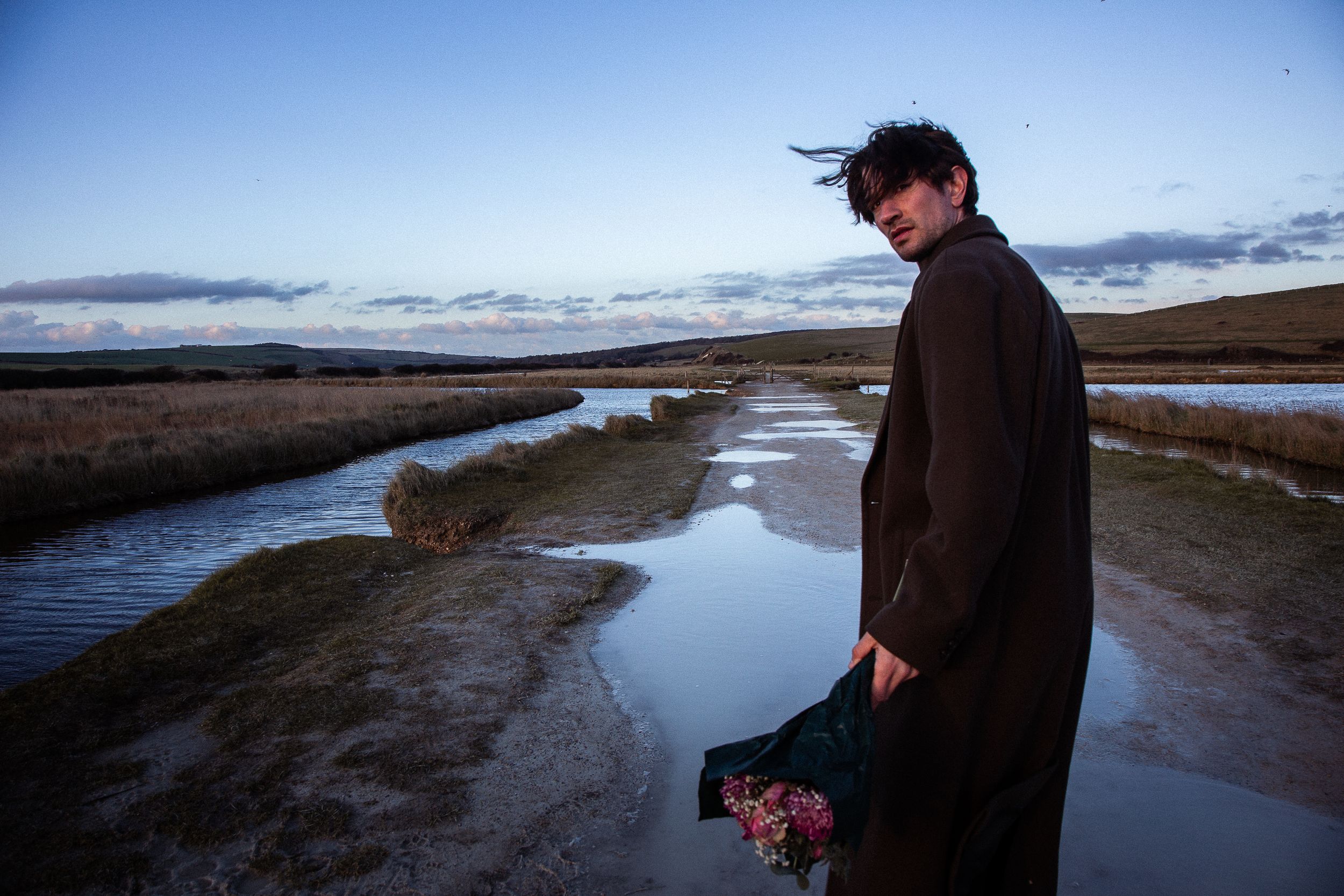 A person in dark clothing stands beside a winding river at dusk, creating a moody silhouette against the water.