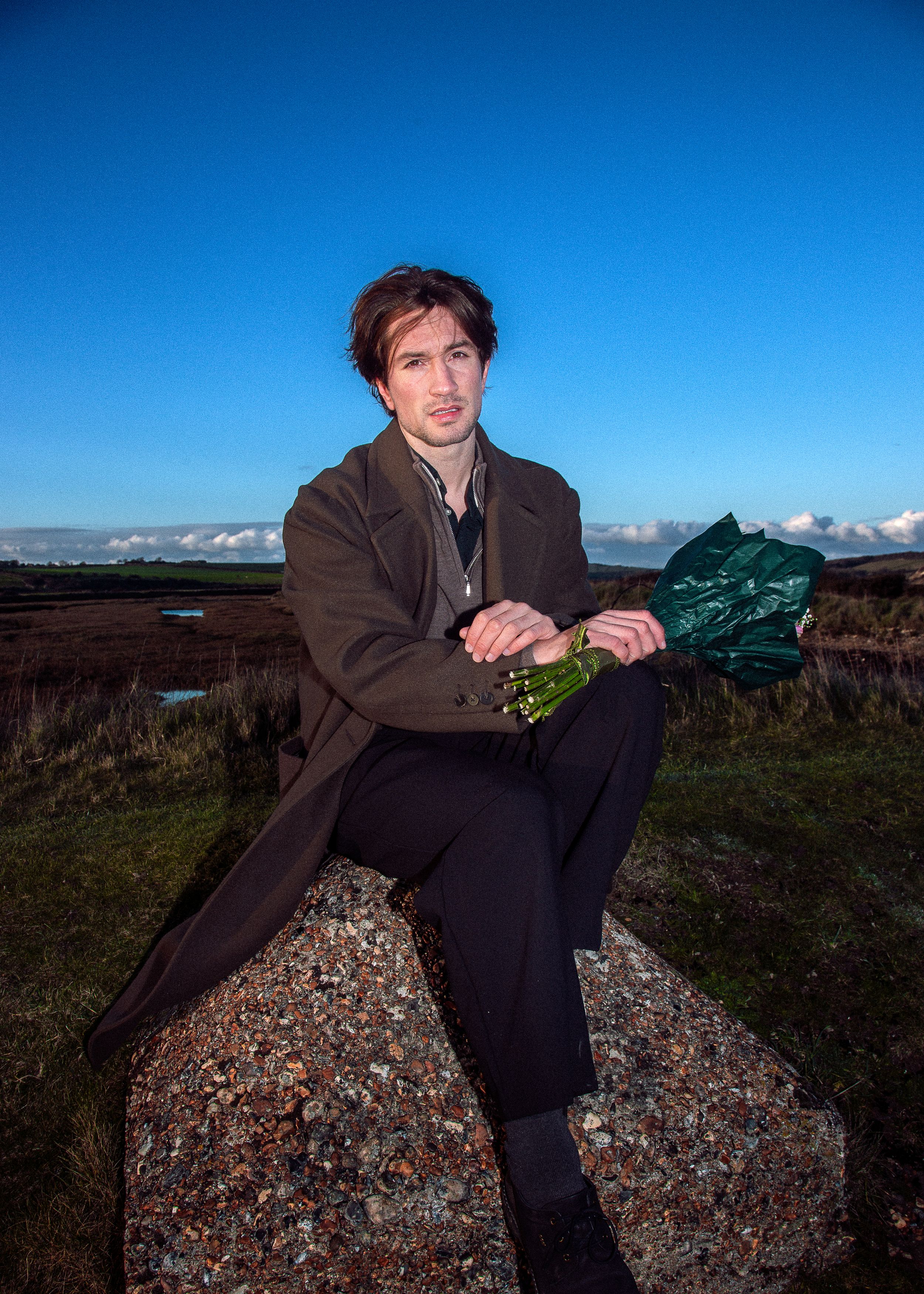 Figure sits on a rocky outcrop against a blue sky wearing dark clothing in a natural outdoor setting.