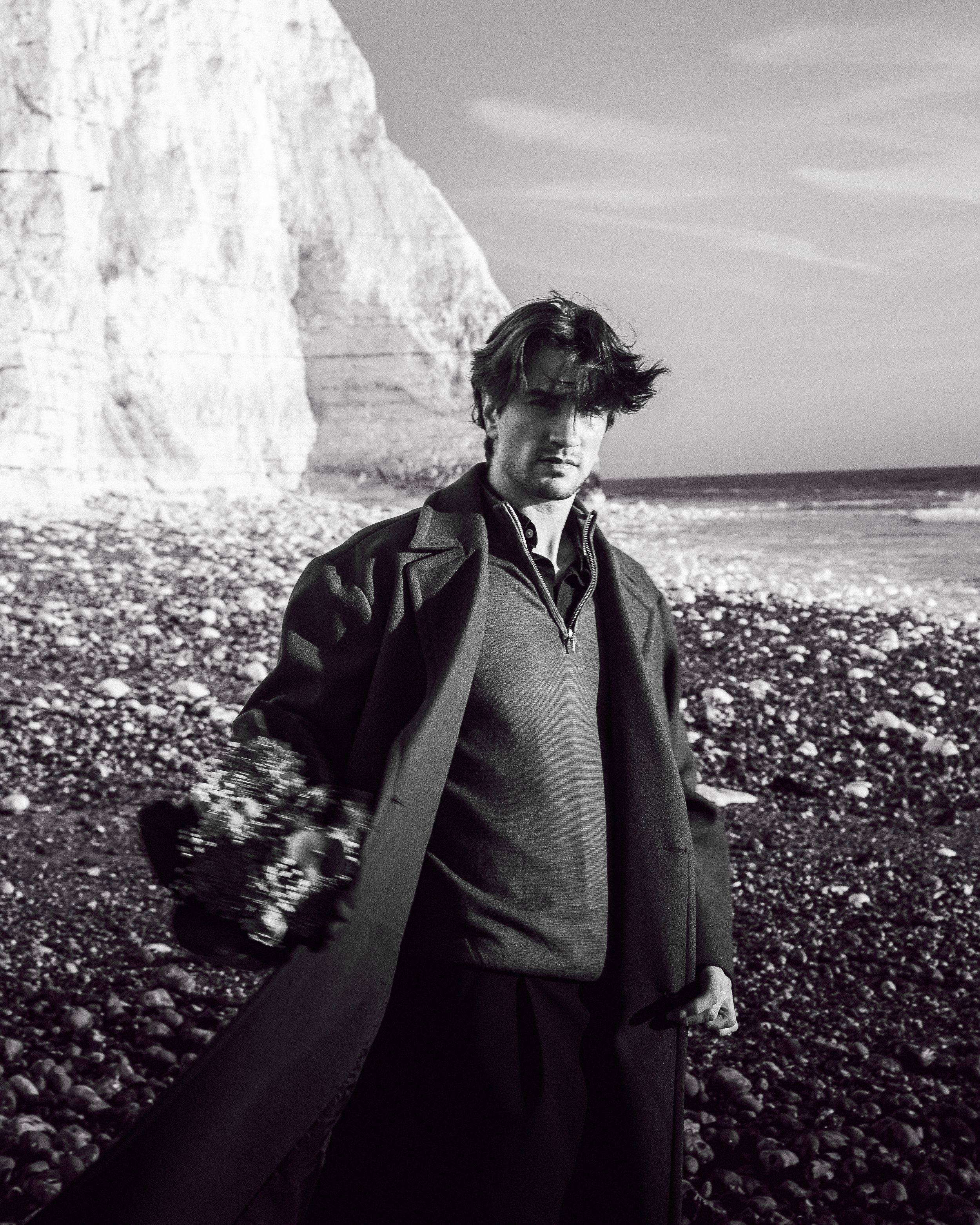 A person wearing dark clothing stands on a rocky beach with white cliffs in the background in black and white.