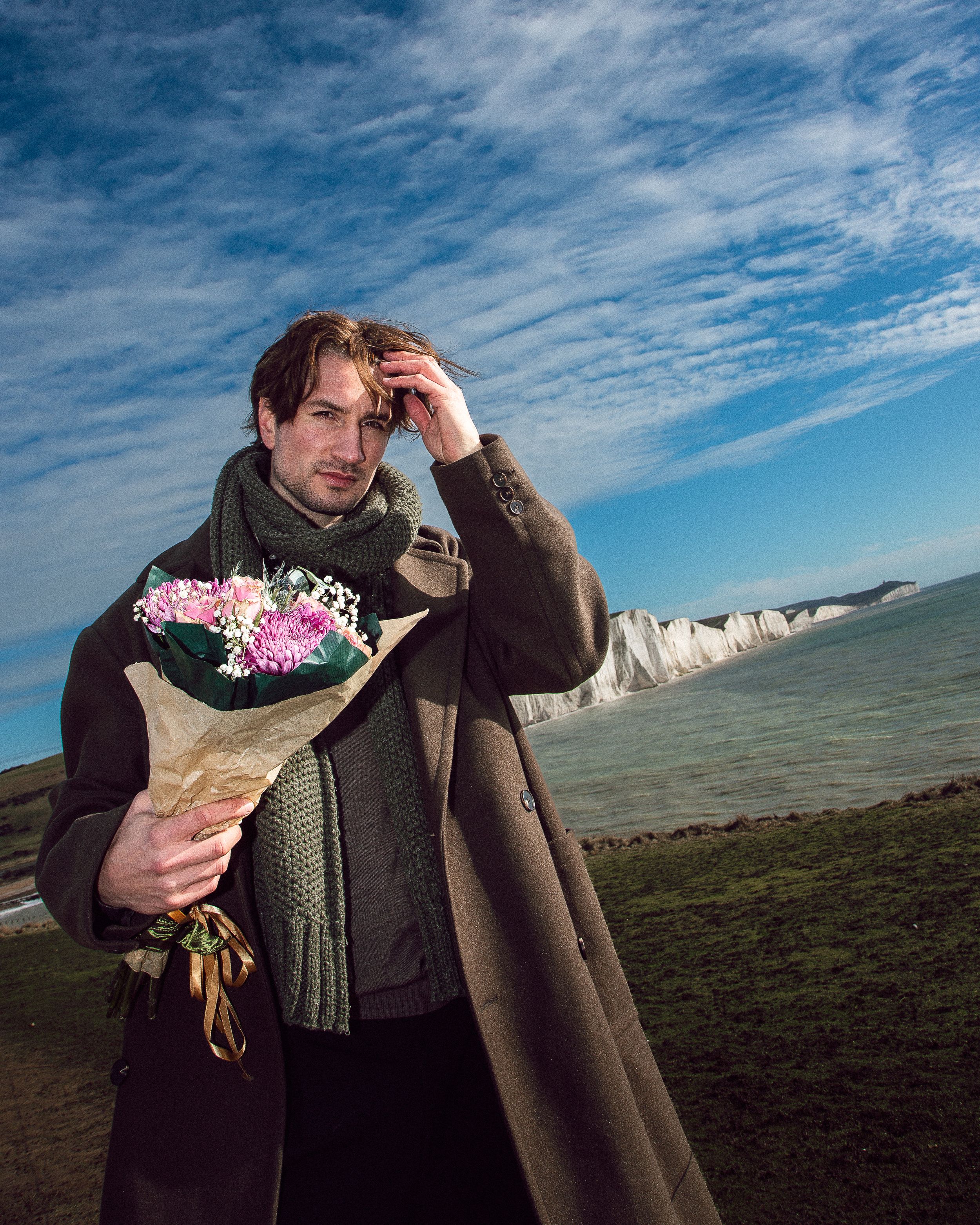 Person holding pink flowers stands against dramatic white cliffs and blue sky with wispy clouds at Seven Sisters coastline.