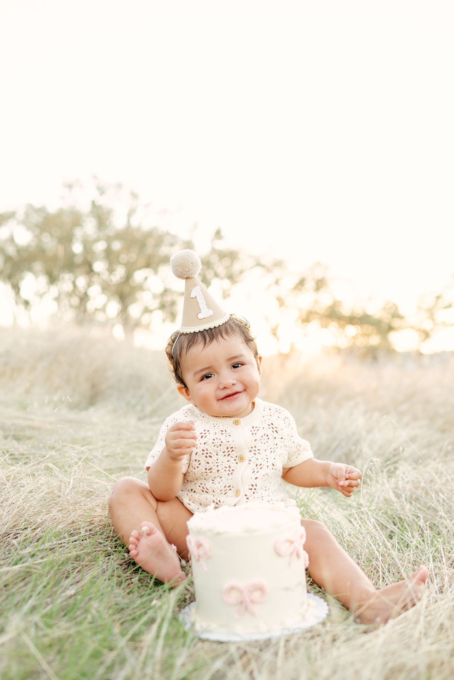 Baby sitting in tall grass during golden hour with pink birthday cake for first birthday cake smash photo session.