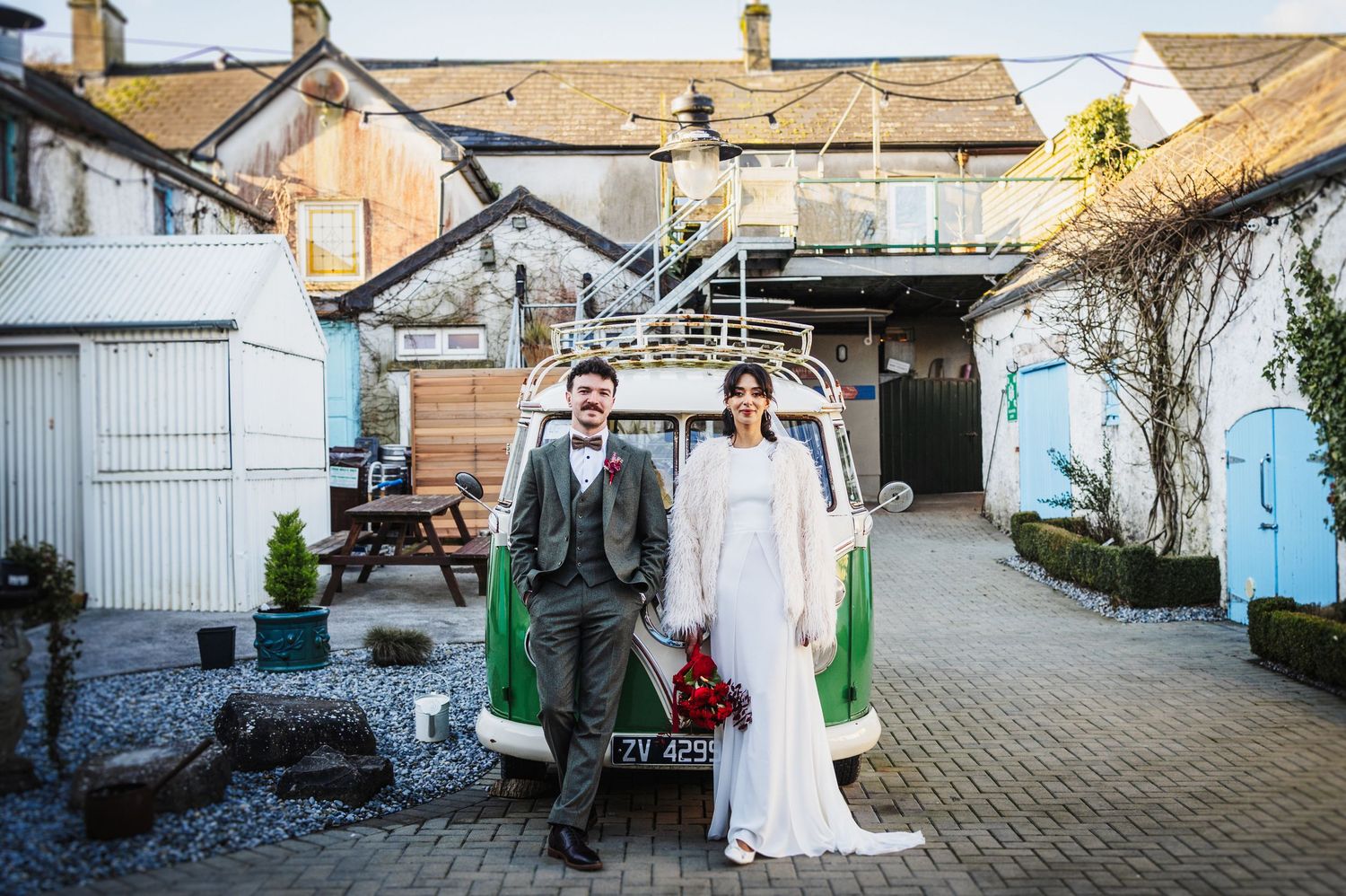 A couple in wedding attire poses in front of a rustic courtyard with stone buildings and blue doors.