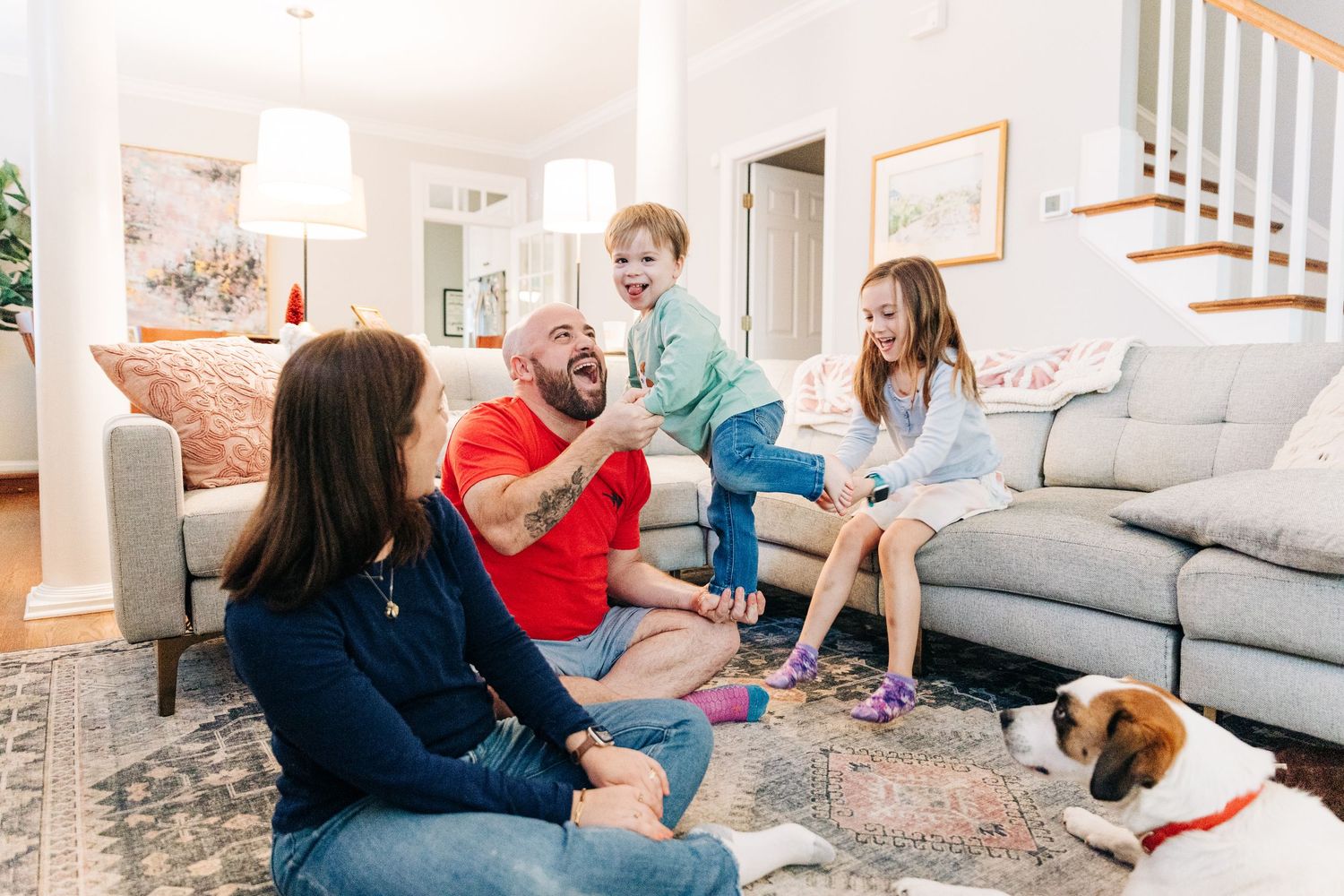 A group sits together on a gray couch in a bright living room while enjoying conversation and laughter as a dad balances toddler on his hand