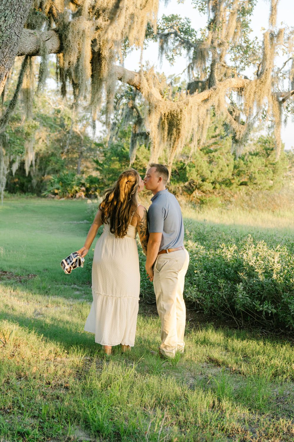 Maternity couples engagement session, woman wearing a white flowy dress while man wears khaki and blue shirt, kissing along the marsh