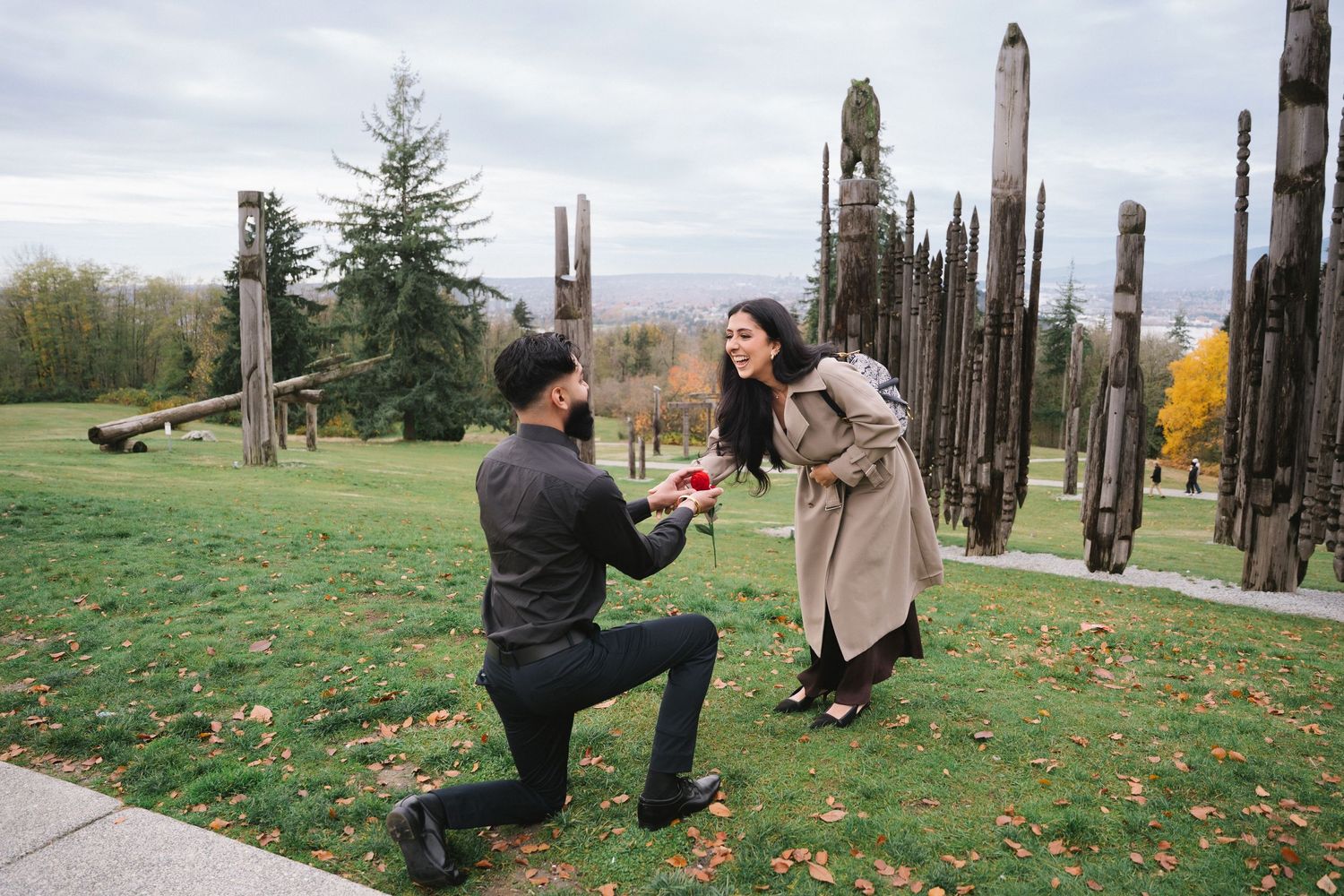 A proposal moment takes place in front of tall wooden totem poles in a park during autumn.