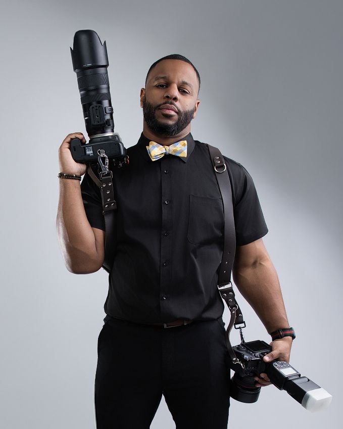 Professional photographer wearing black attire poses with camera equipment against gray studio backdrop.