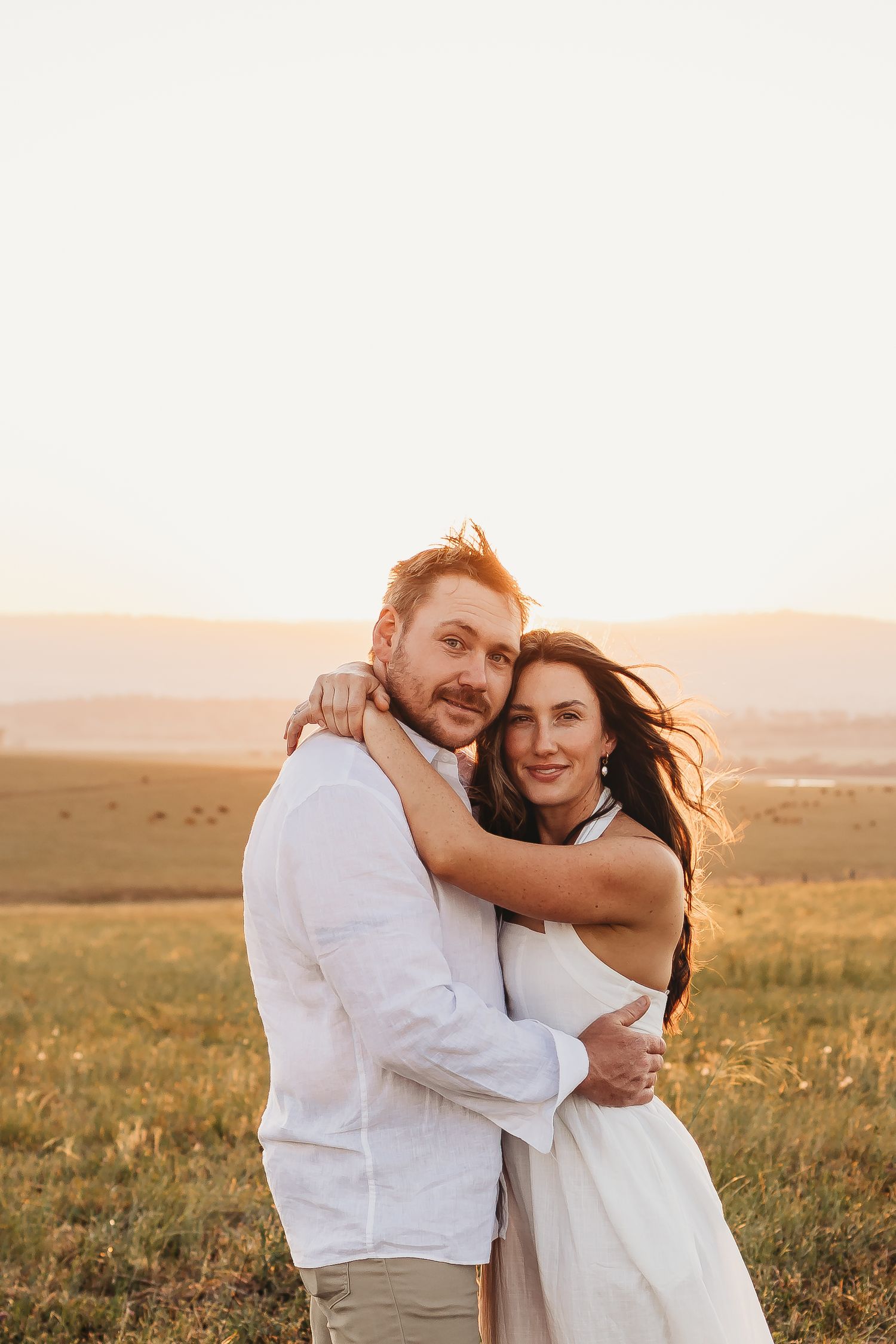A couple embraces in a sun-drenched meadow during golden hour, wearing white attire against a scenic countryside backdrop.