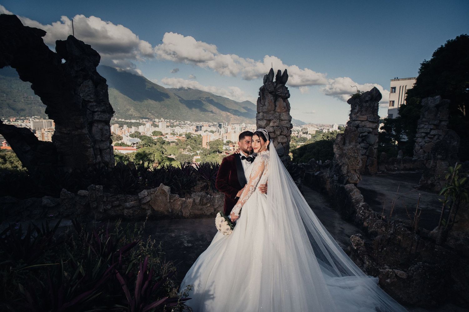 A couple in wedding attire embraces at ancient castle ruins overlooking a scenic mountain valley landscape.