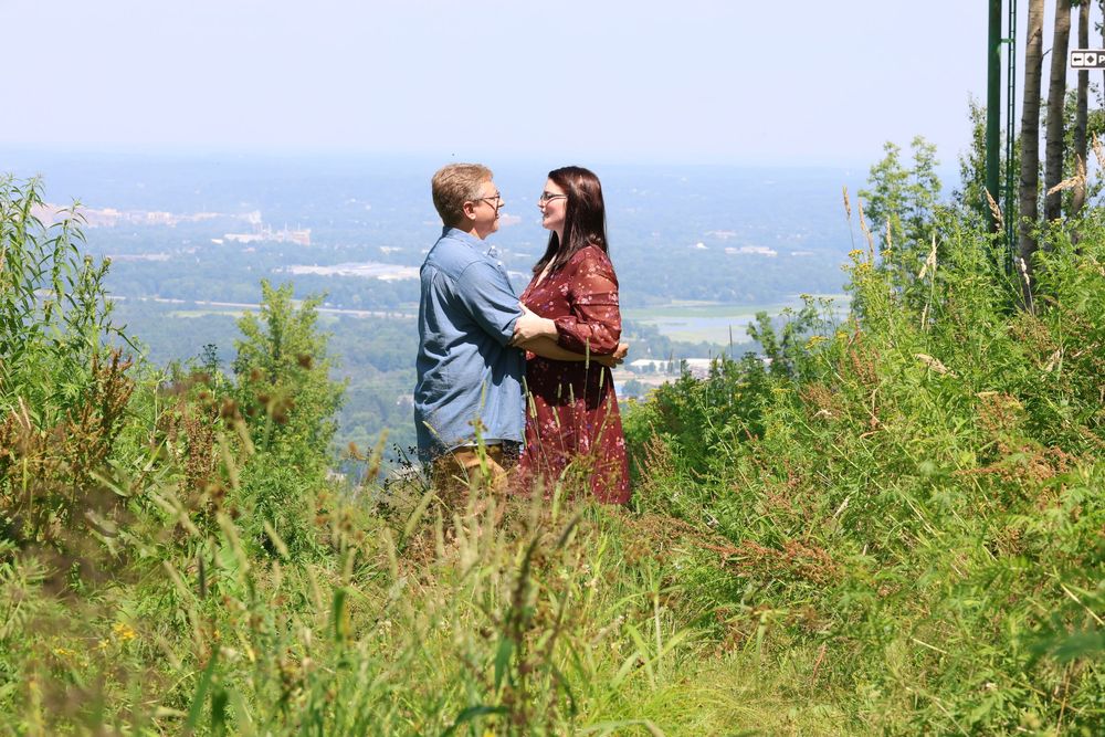 Two people stand on a hilltop overlooking a scenic valley landscape on a sunny summer day.