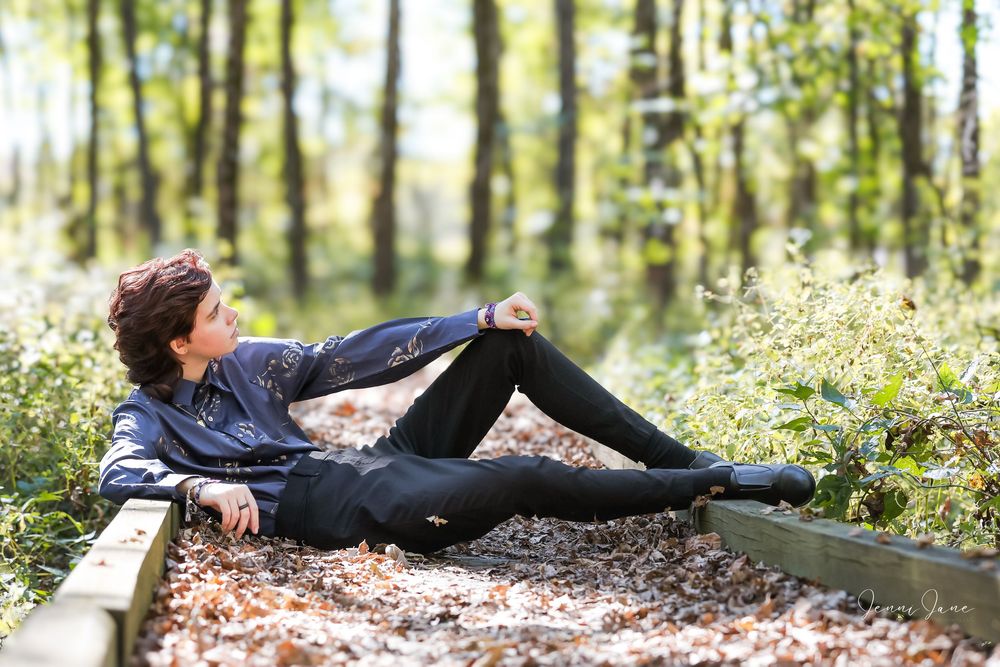 Person in casual attire relaxing on wooden boardwalk surrounded by lush forest and wildflowers.