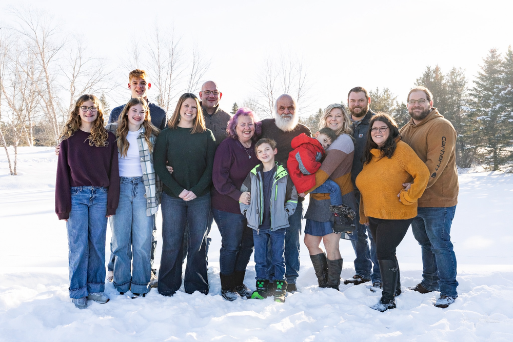 A group of people stand together posing in a snowy winter landscape with evergreen trees in the background.