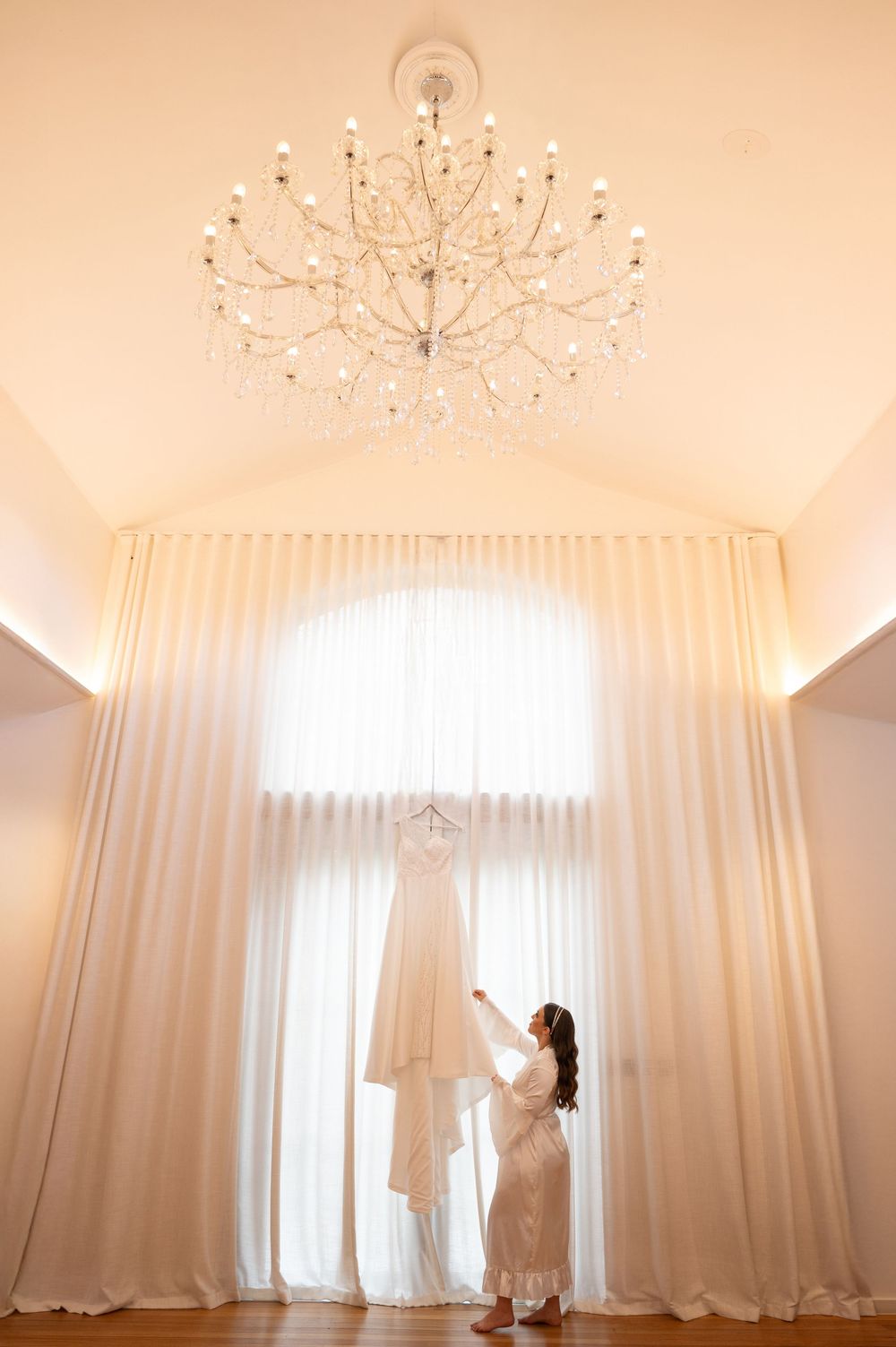 Bride in white robe reaching for her wedding gown hanging by a grand arched window beneath a crystal chandelier.