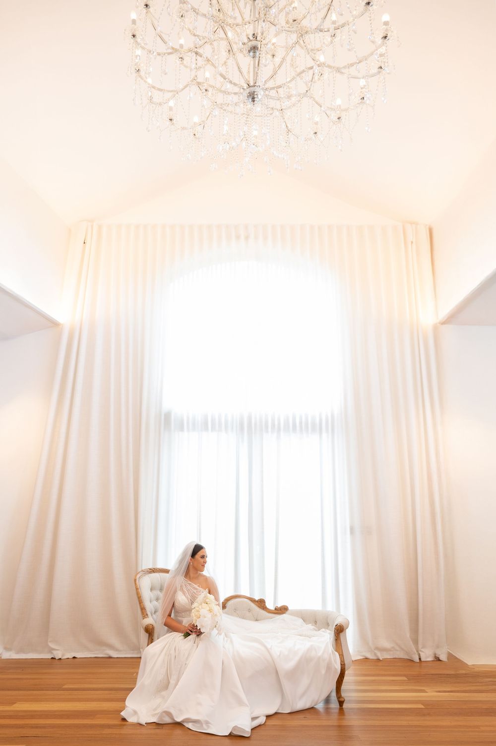 Bride in white ball gown seated alone on vintage sofa beneath crystal chandelier in bright elegant bridal suite.