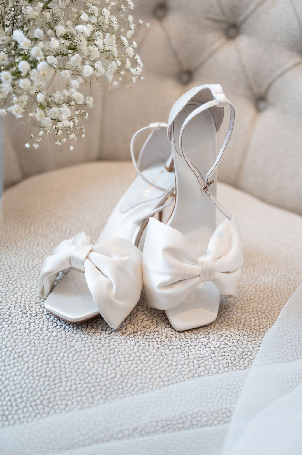White satin bridal heels with bow detail displayed on cream upholstered chair.