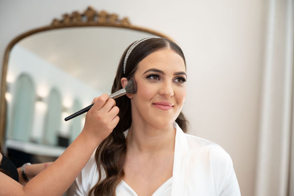 Bride in white robe smiles while makeup artist applies blush before wedding ceremony.