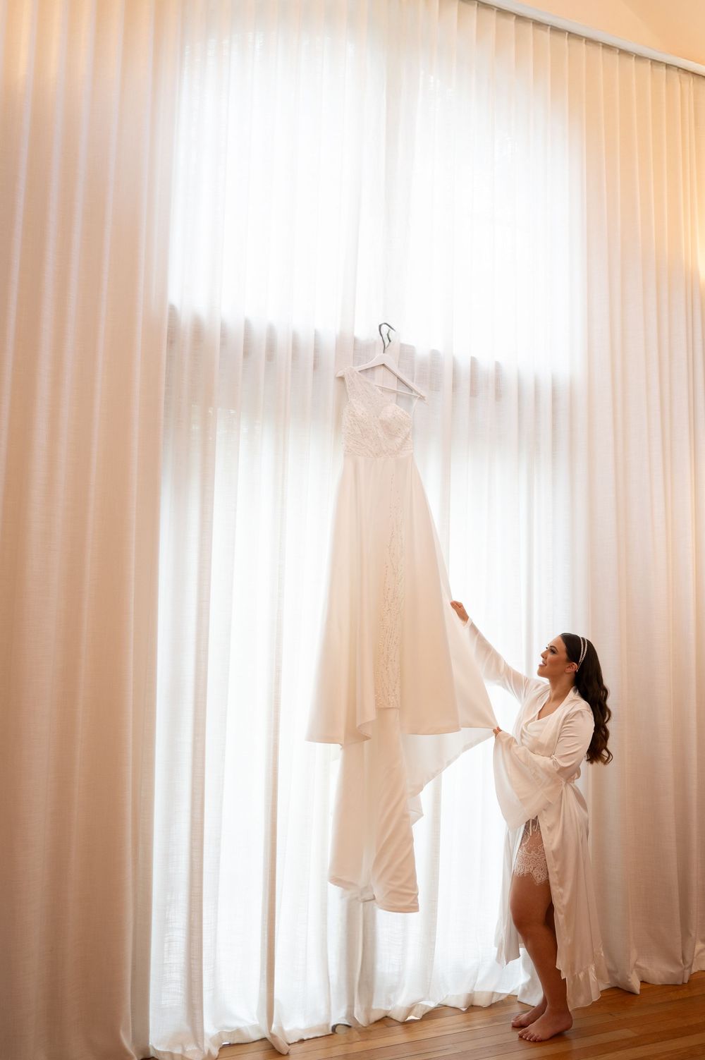 Bride in white robe reaches for wedding dress hanging in sunlit window.