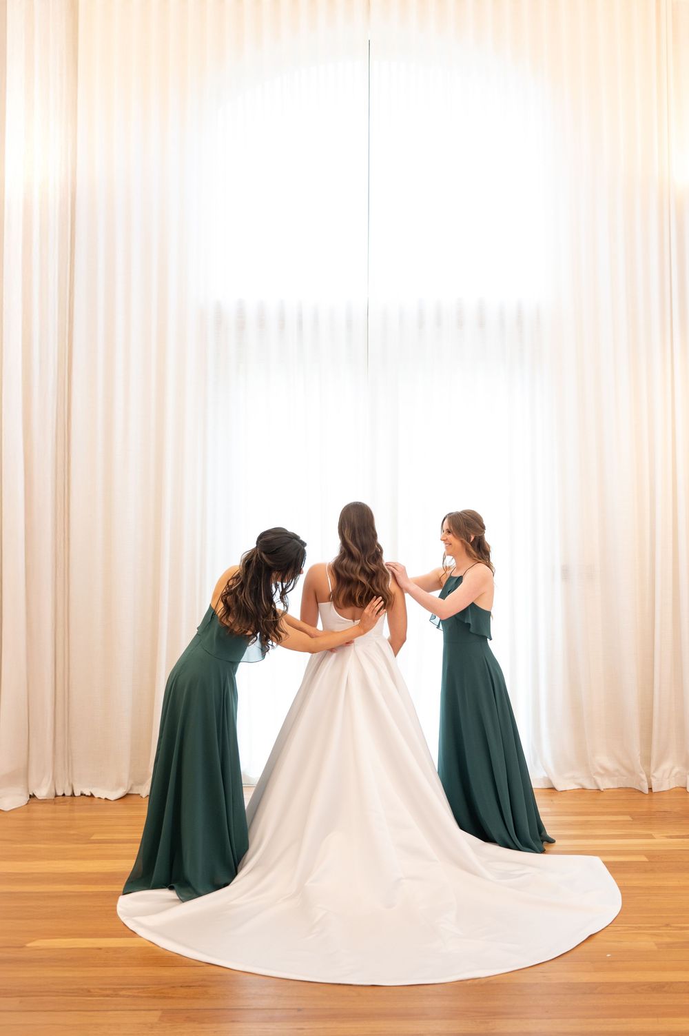 Two bridesmaids in emerald green gowns helping bride into her white ball gown by a large arched window.