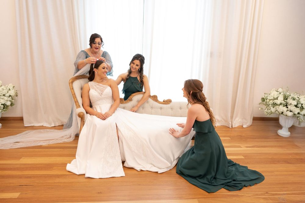 Bride in white gown seated on vintage sofa surrounded by bridesmaids in green dresses in elegant bridal suite.