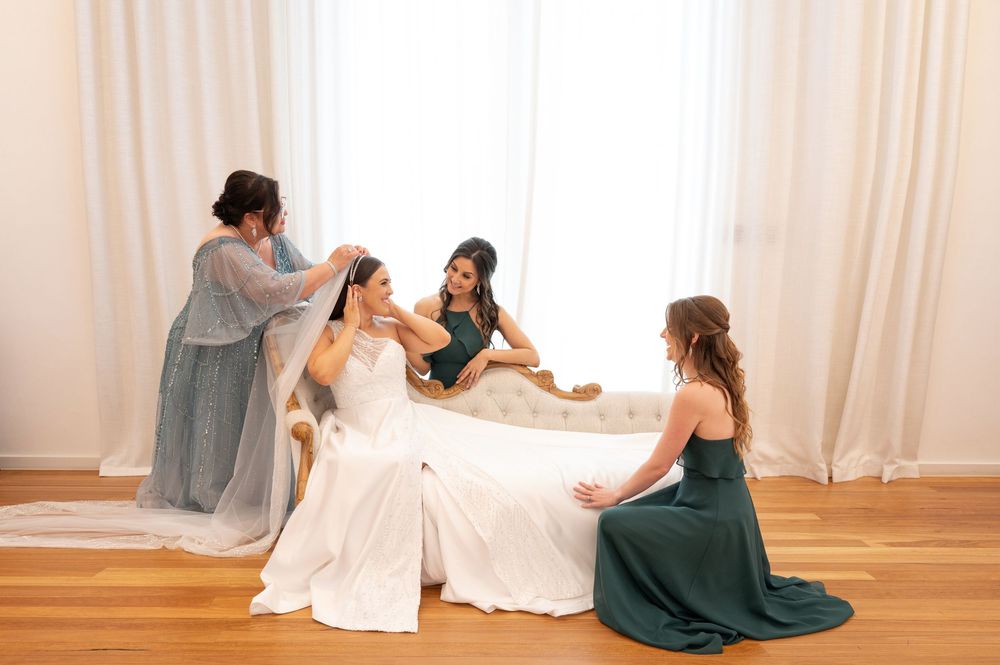 Bridesmaids in green and blue gowns helping bride with veil while seated on ornate sofa by bright window.