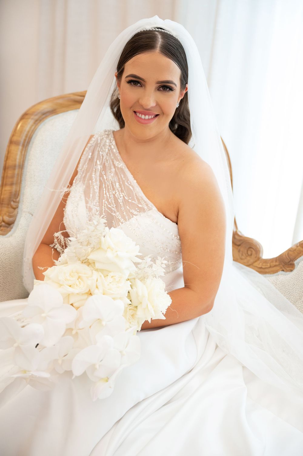 Radiant bride in one-shoulder lace gown holding white bouquet while seated on gold vintage chair with veil.