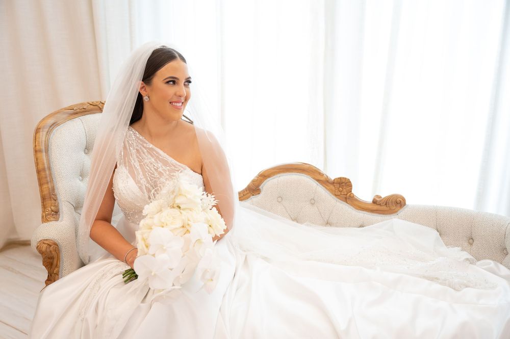 Elegant bride in one-shoulder wedding gown holding white floral bouquet while seated on ornate vintage sofa.