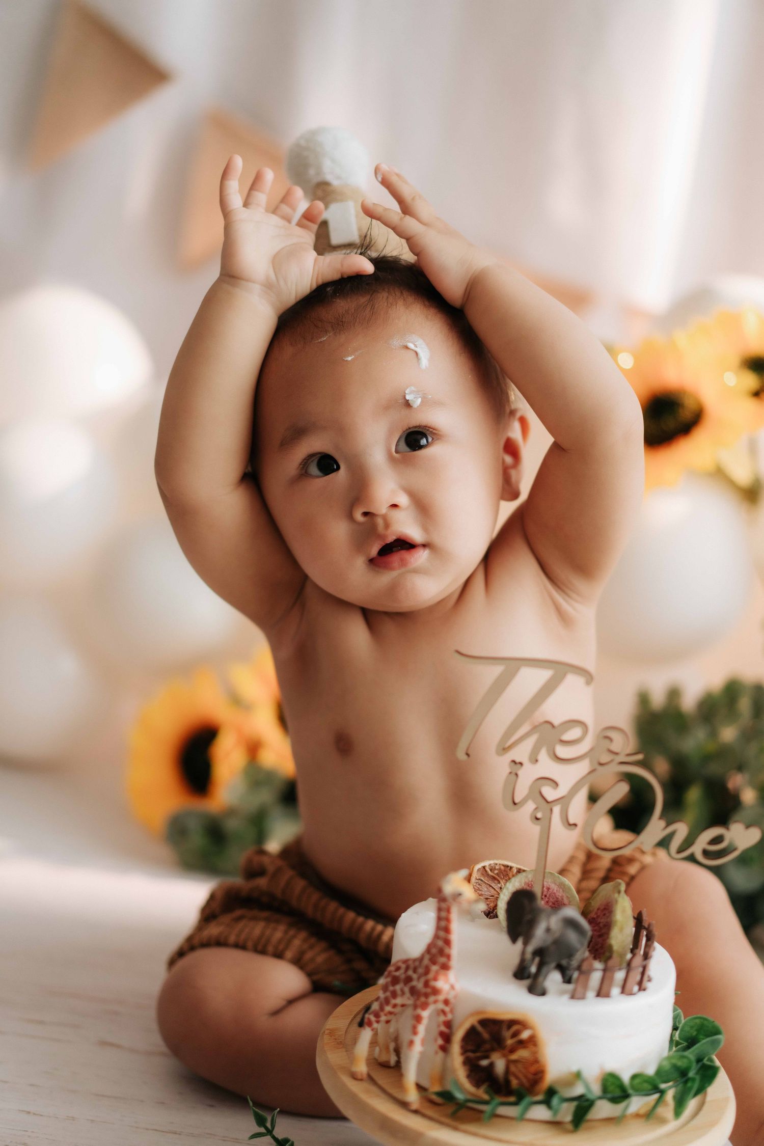 Baby in brown diaper sits with cake and toy animals during first birthday photoshoot with sunflowers in background.