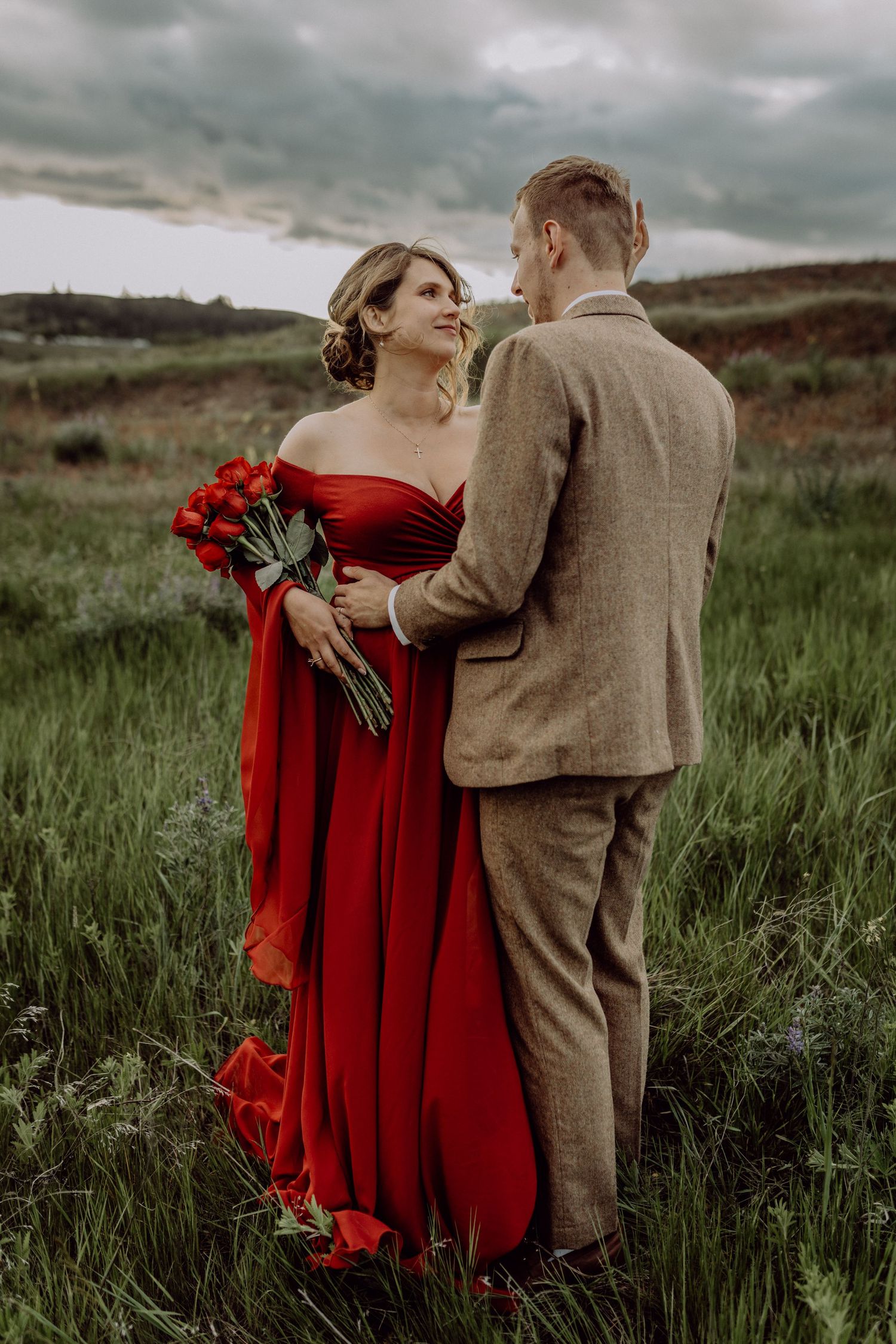 A romantic couple embraces in a grassy field, with the woman wearing a flowing red dress and holding red flowers.