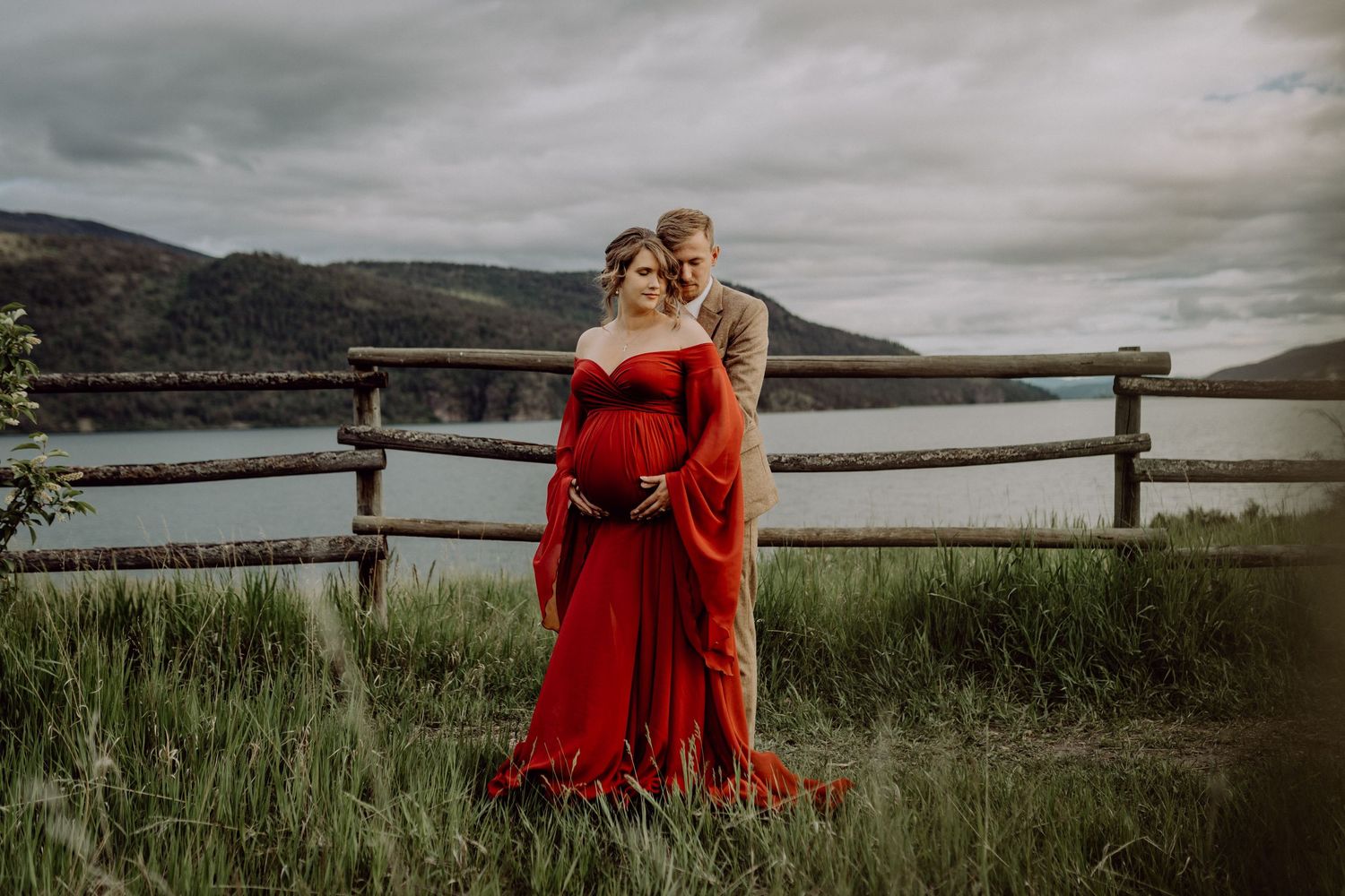 A person in a flowing red dress poses gracefully in a scenic mountain landscape near a lake at dusk.