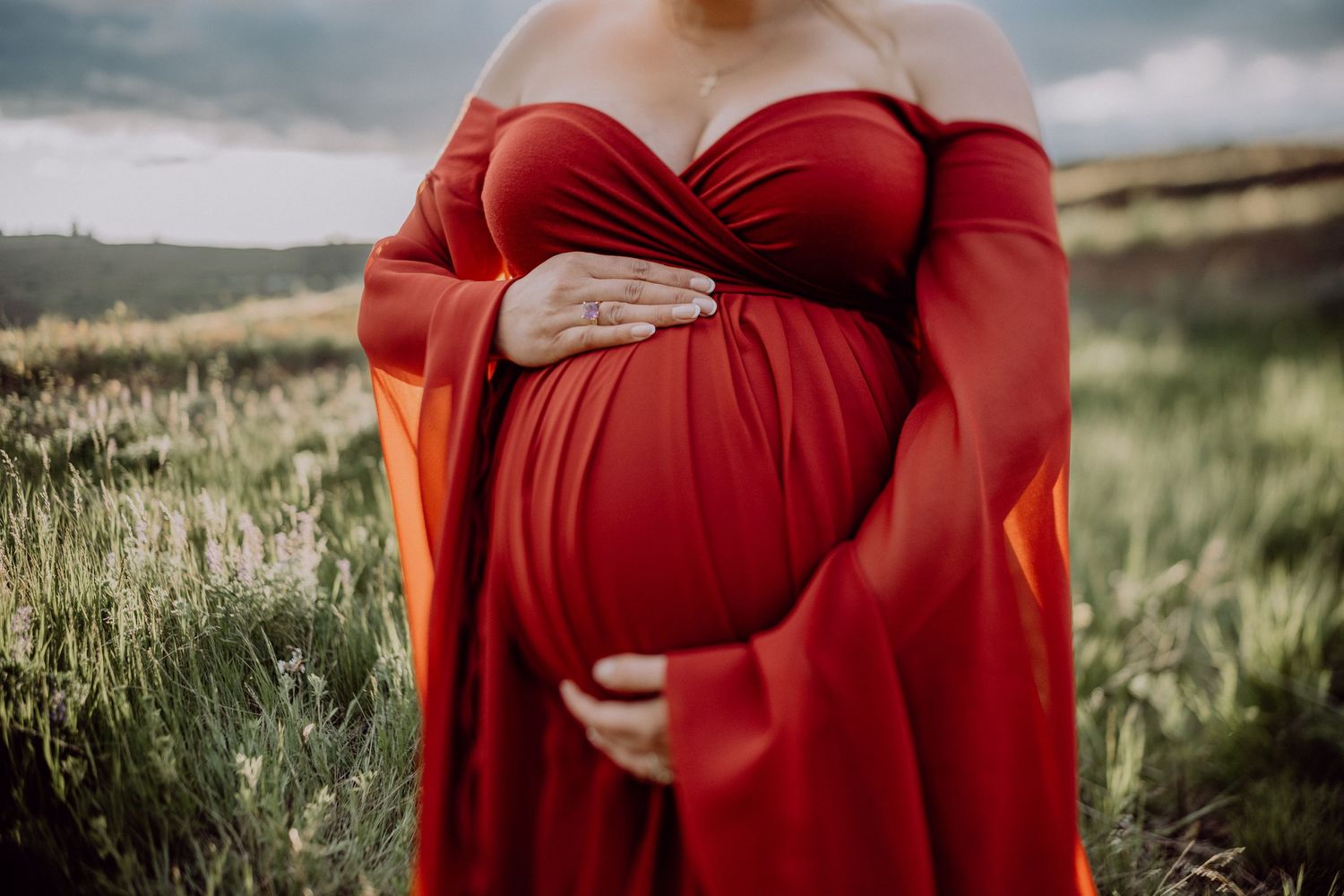 A pregnant woman in a flowing red dress cradles her belly while standing in a grassy field at sunset.