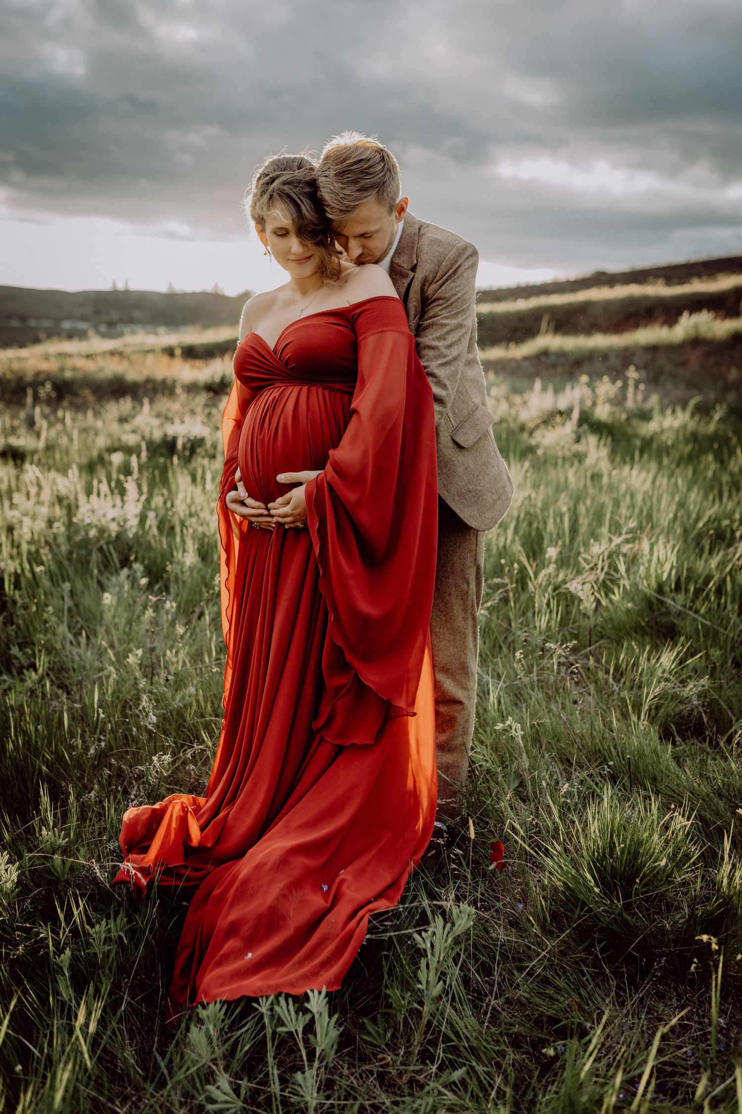 A couple embraces in a grassy field under moody skies, with the woman wearing a flowing red dress.