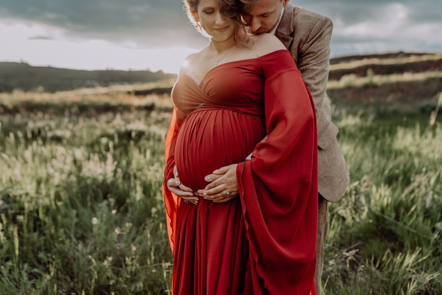 A couple embraces in a grassy field wearing a flowing red dress and neutral tones at sunset.