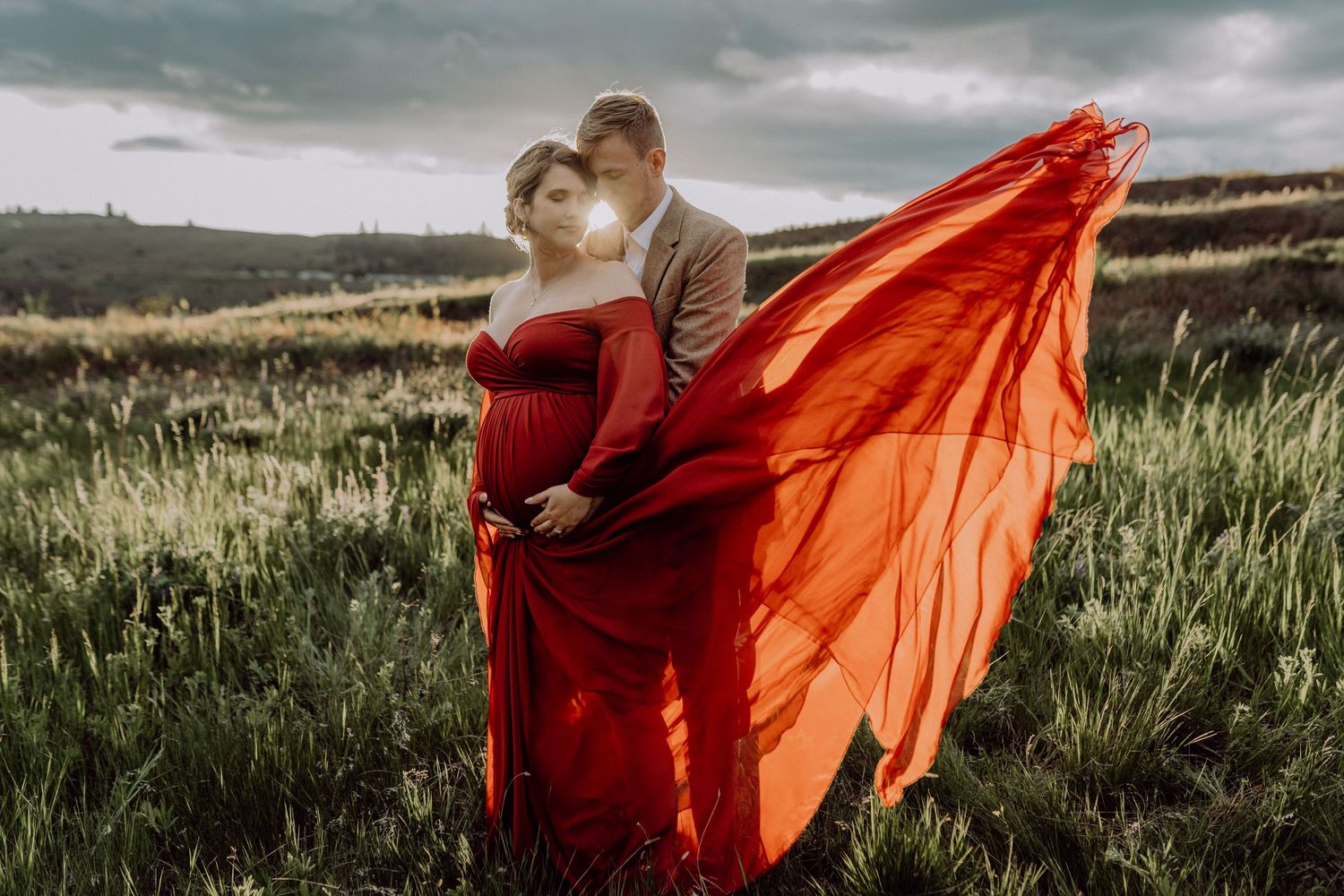A couple embraces in a dramatic field landscape while an orange fabric billows in the wind against moody storm clouds.