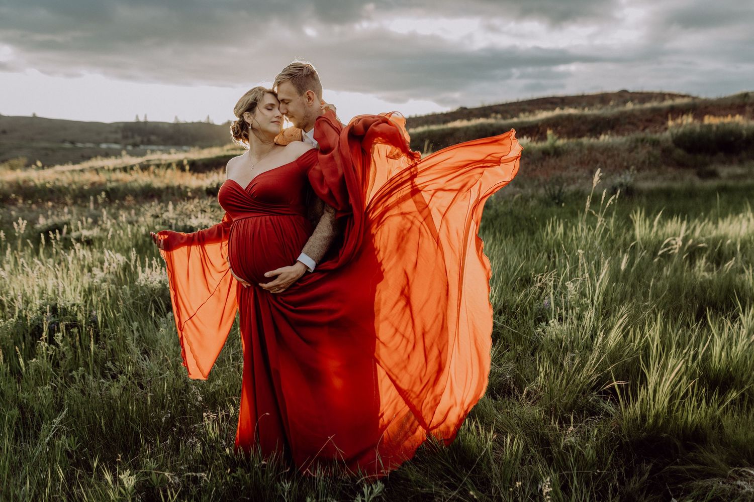 Dancers in flowing red and orange dresses move gracefully through a green meadow landscape under moody skies.