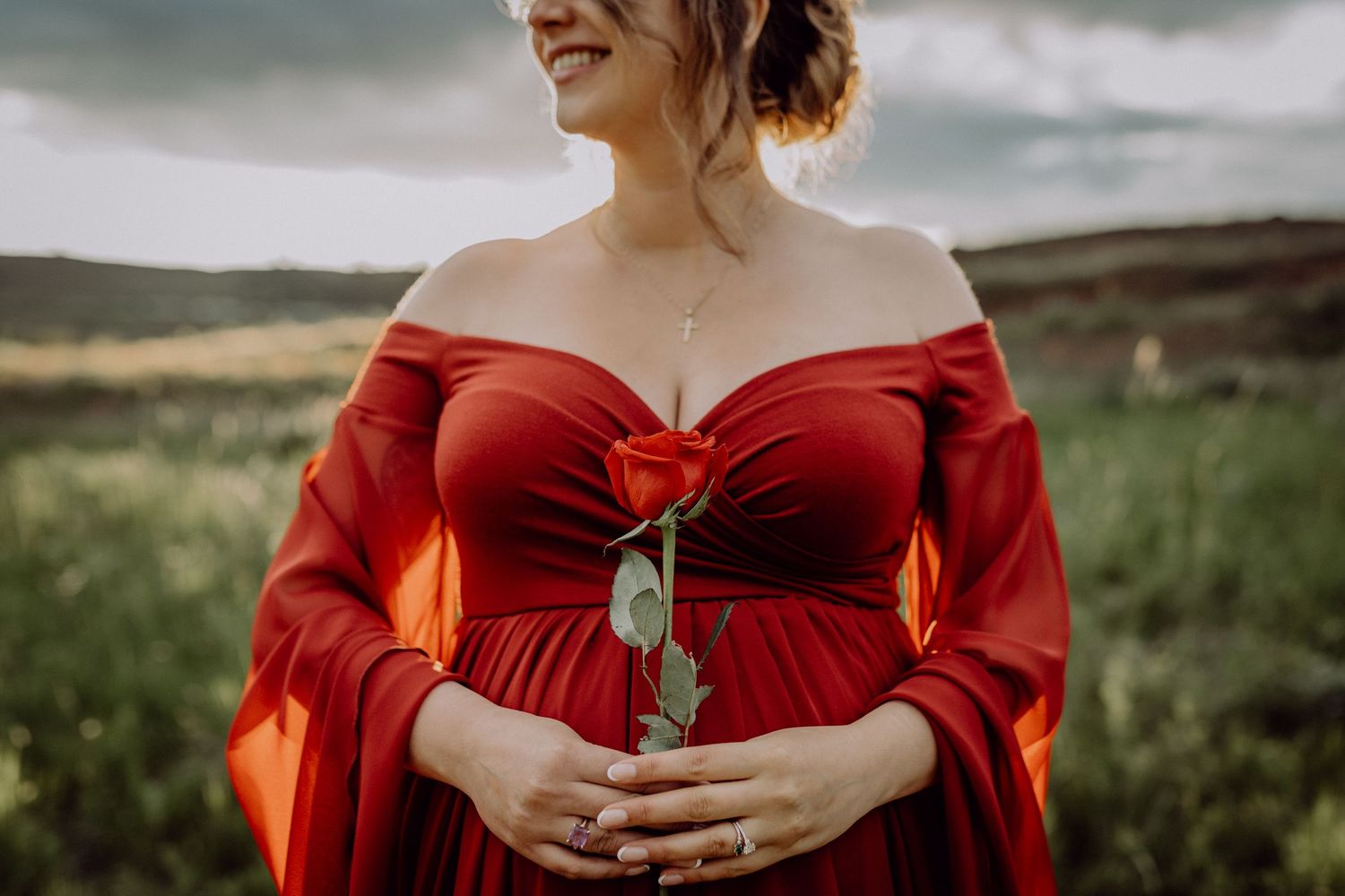 A person in a flowing red off-shoulder dress poses in a scenic green meadow landscape during golden hour.