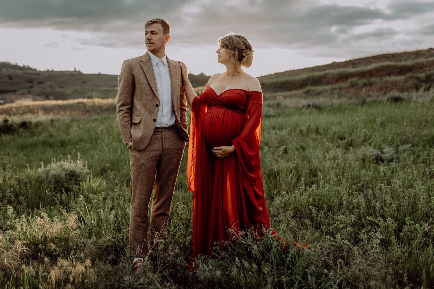 A couple in formal attire embraces on a scenic hilltop meadow during golden hour.