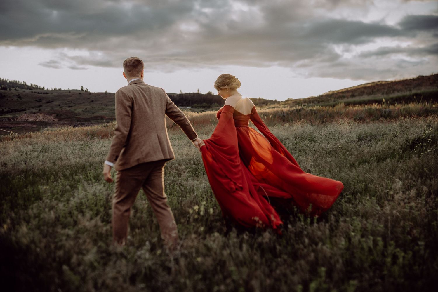 A couple stands in a windswept field at dusk, with a flowing red dress billowing dramatically against the moody sky.