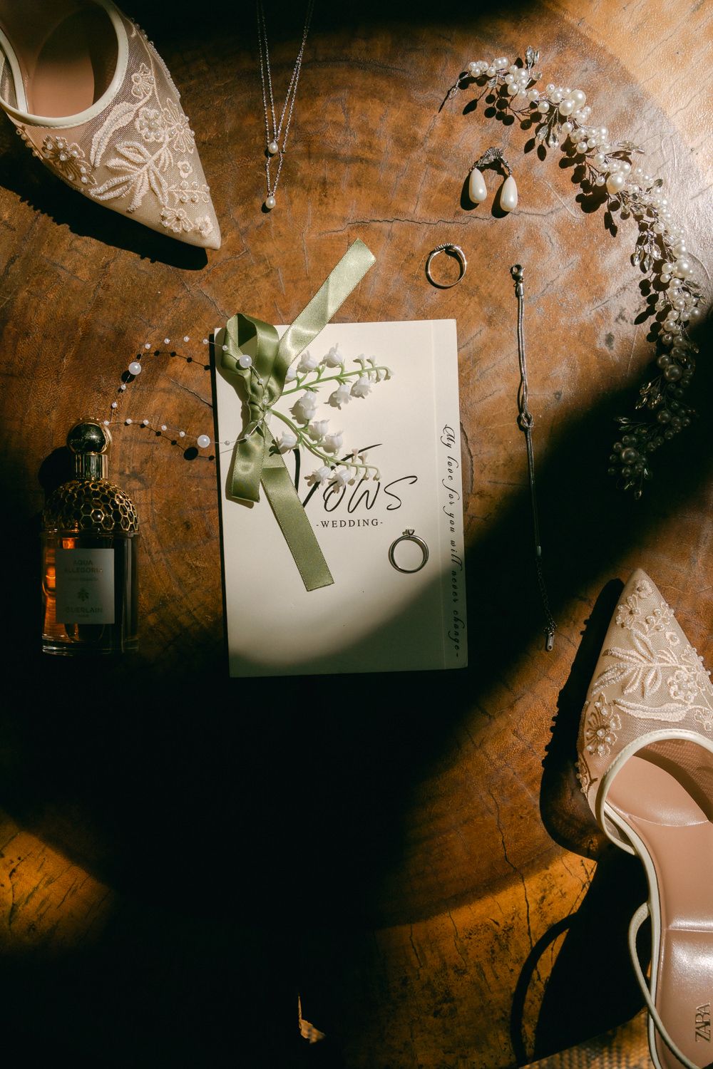 Wedding vows card with delicate lace shoes, dried flowers, and jewelry arranged on wooden surface in moody lighting.