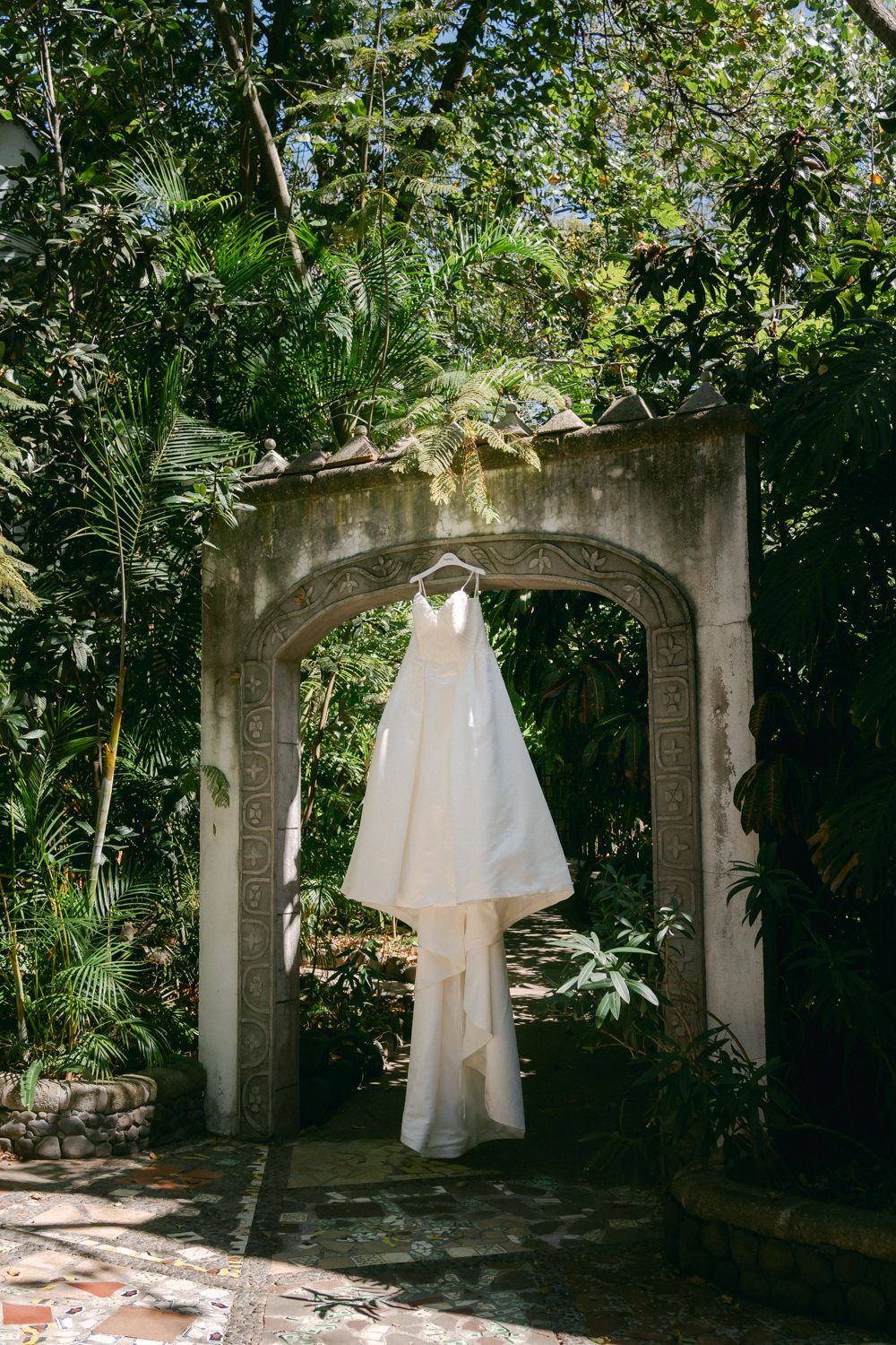 A white wedding dress hangs in an outdoor garden setting with stone archways and lush greenery.