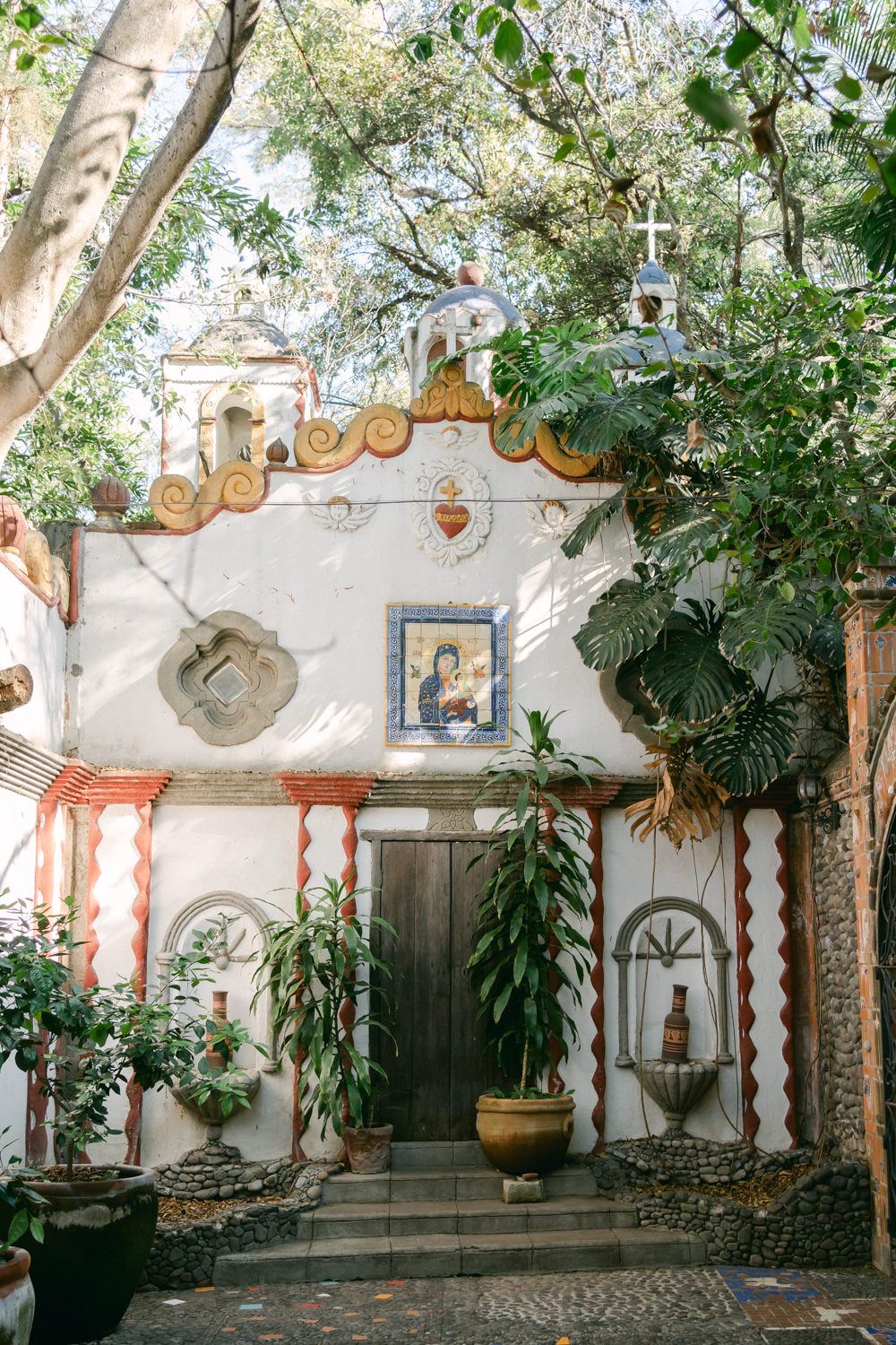 Charming Mediterranean-style entryway with ornate white walls, wooden door, and hanging lanterns amid lush garden foliage.