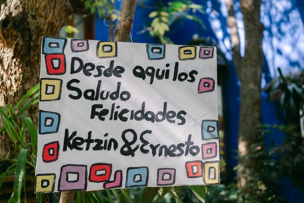 A handwritten Spanish greeting sign with colorful doodles and decorative borders hangs on a tree.