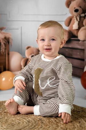 Baby in striped onesie sits on jute rug with teddy bears and balloons in background.