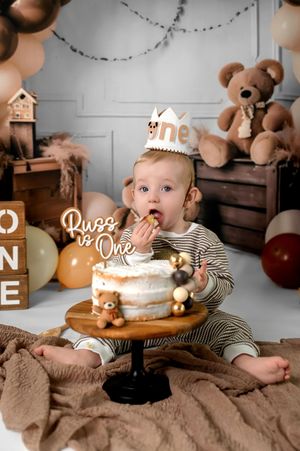 Small figure in striped outfit sits on brown blanket surrounded by teddy bears and wooden crates in studio setting.