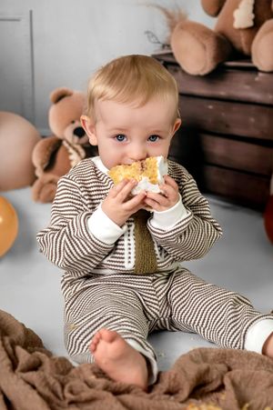 Figure in brown and white striped outfit enjoys snack while sitting on cozy blanket with teddy bears in background.