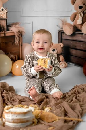 A toddler in striped clothing sits on a couch with brown blankets and stuffed animals while eating a snack.