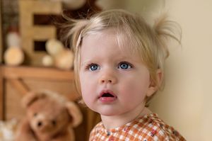 A toddler with bright blue eyes and blonde hair gazes upward while sitting indoors.