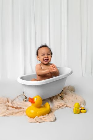 Baby sitting in white bathtub with rubber ducks on beige fabric against white curtain backdrop.